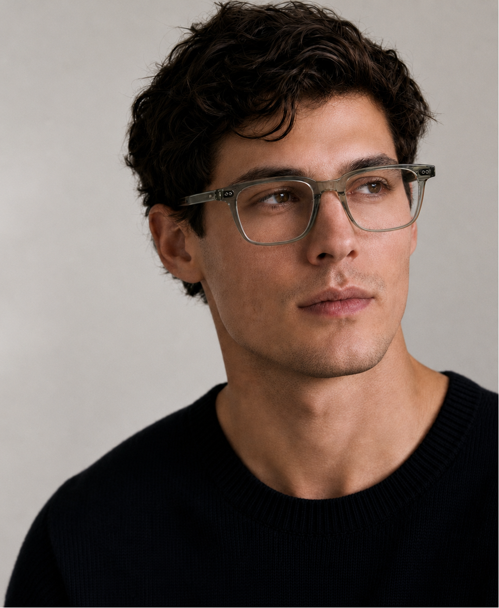A close-up portrait of a man wearing square blue light glasses and a dark crewneck sweater against a soft light gray background.